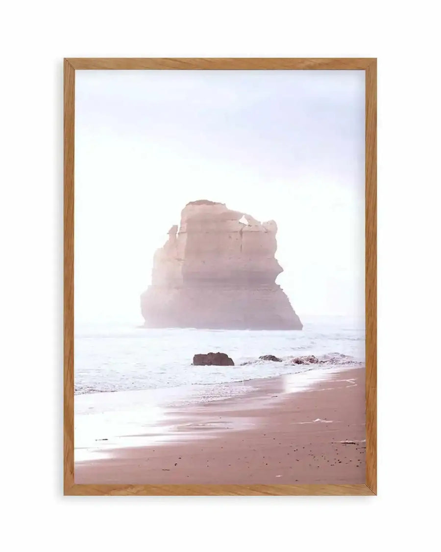 Framed photography art print of a coastal scene at dusk with a natural oak frame and white border. A large, light pink rock formation stands in the ocean, with gentle waves breaking on a wet, sandy beach in the foreground.