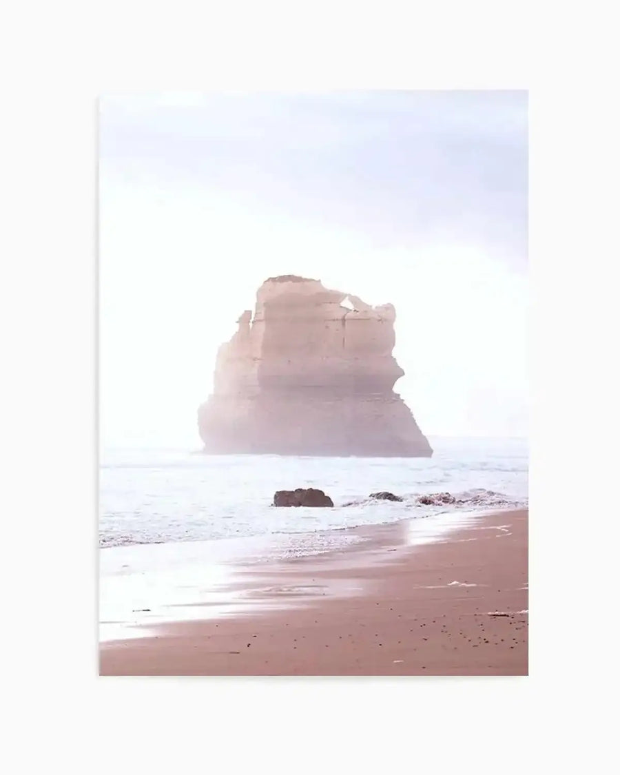 Photography art print of a coastal beach scene at dusk, featuring a large, light pink rock formation in the distance, surrounded by gentle white waves. The foreground shows wet, sandy beach reflecting the pale sky, with smaller dark rocks near the water's edge.