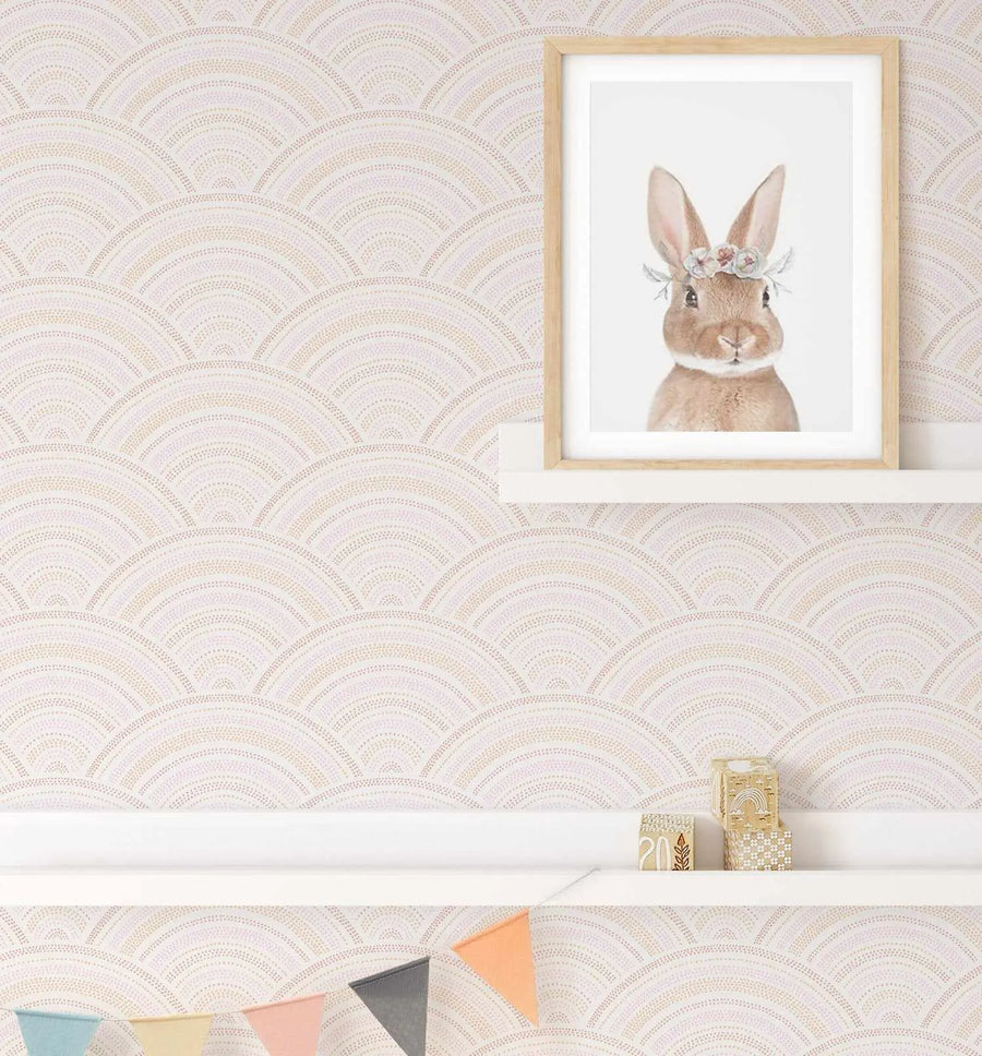 Lifestyle shot featuring Earth Rainbows Wallpaper in a child's room. The wallpaper has a repeating scallop pattern made of dotted lines in soft pink, cream, and warm gold tones, creating a gentle, textured effect. A white floating shelf is mounted on the wall, holding a framed art print of a brown bunny wearing a floral crown. Below, another white shelf displays a colourful bunting with flags in pastel blue, yellow, pink, and grey.