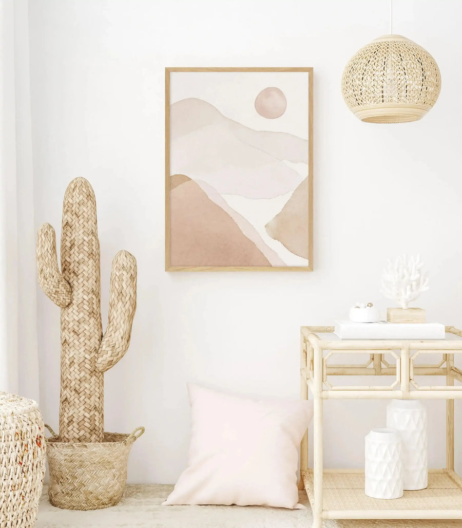 Lifestyle shot of a minimalist room featuring an art print with abstract, layered mountain shapes in various shades of dusty rose and pale peach, with a circular sun-like shape in the upper right, displayed in a natural wood frame. The room includes a large woven straw cactus, a light pink cushion, a rattan side table with white decorative items, and a woven pendant light.