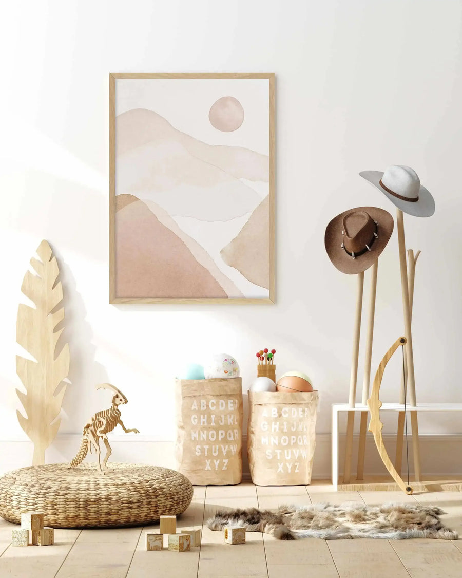 Kids Room:
Framed art print displayed on a white wall in a neutral-toned kids' room. The print features an abstract watercolour landscape with layered, organic shapes in shades of dusty rose, beige, and cream, with a pale pink sun in the upper right, and a natural wood frame. The room includes a large wooden feather, a woven pouf, a wooden dinosaur skeleton, alphabet storage bags, a wooden coat rack with two cowboy hats, a small wooden bow, and a faux fur rug.
