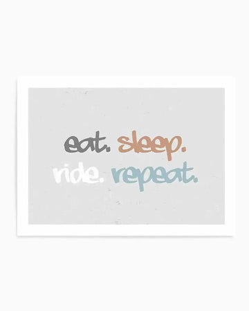Art print featuring the phrase "eat. sleep. ride. repeat." in a casual, handwritten-style font on a light grey background with a subtle textured effect. "eat." is in dark grey, "sleep." is in a muted terracotta orange, "ride." is in light grey, and "repeat." is in a dusty blue. The print is displayed in a thin white frame.
