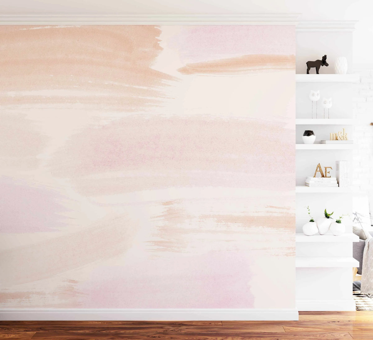 Lifestyle shot of a living room featuring Elani Abstract Wallpaper on the left wall. The wallpaper features large-scale abstract brushstrokes in soft blush pink, pale peach, and cream, creating a gentle, ethereal pattern. The room has light wood flooring and white baseboards. To the right, a white built-in shelving unit displays various decorative items, including a black moose figurine, white vases, and books.