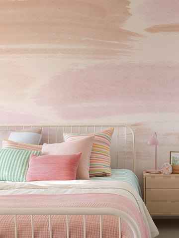 "Peel and stick wallpaper featuring an abstract watercolor design with broad, horizontal brushstrokes in varying shades of blush pink, dusty rose, and pale cream, creating a soft, cloud-like effect. The wallpaper is displayed on the wall behind a white metal bed frame with multiple pillows in shades of pink, mint green, and multicolored stripes. A light wood bedside table with a pink lamp and a small clock is visible on the right."