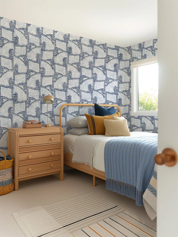 A bedroom featuring a wallpaper with a repeating pattern of abstract, organic shapes in shades of deep indigo blue on a cream background. The design has a hand-drawn, block-printed quality with visible texture. The wallpaper covers the wall behind a light wood bed frame with a blue and yellow patterned throw blanket, and a matching light wood nightstand with a gold lamp.
