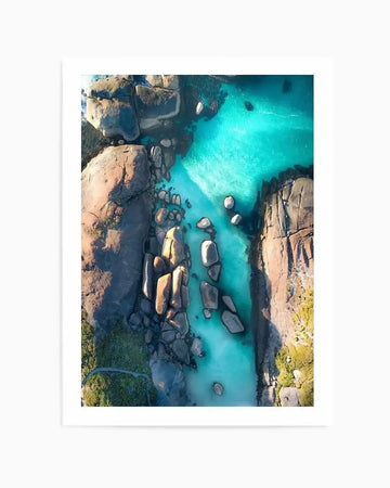 Framed aerial photograph of a vibrant turquoise ocean channel winding between large, smooth granite boulders and rocky shorelines, with a natural wood frame and white border. The clear water reveals sandy patches and submerged rocks, while the land features sparse green vegetation.