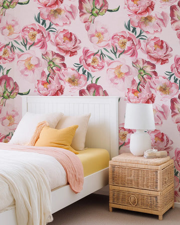 "Peel and stick wallpaper featuring a repeating pattern of large-scale pink peony blooms with soft brushstroke detail and green leaves on a light pink background, displayed on a bedroom wall above a white bed with yellow sheets, white duvet, and a pink throw. A white lamp sits on a woven rattan bedside table."