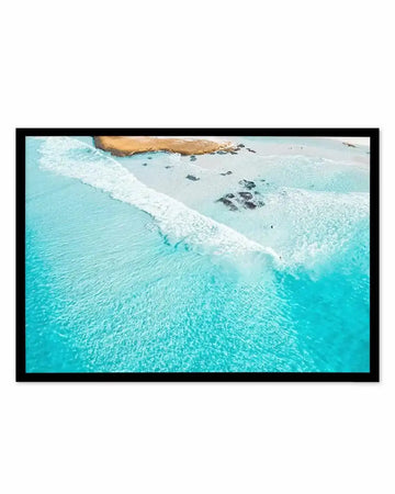Aerial photograph of turquoise ocean waves rolling onto a sandy beach with dark rocky outcrops, captured from above showing gradient from bright aqua water to white foam to pale sand, framed in black with white border.