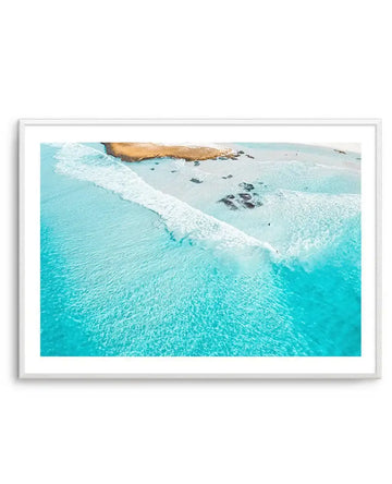 Aerial photograph of a turquoise ocean with white wave foam rolling toward a sandy beach dotted with dark rocks. Golden-brown rocky outcrops visible at the top. The image is framed in a silver-toned frame with a white border, creating a horizontal landscape composition.