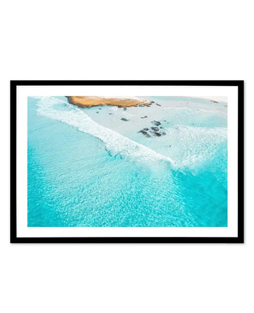 Aerial photograph of a turquoise ocean with white-capped waves rolling toward a sandy beach dotted with dark rocks. Golden-brown rocky outcrops visible at the top of the frame. The image is mounted in a black frame with a white border, displaying the coastal landscape with rich turquoise, cream, and tan tones.