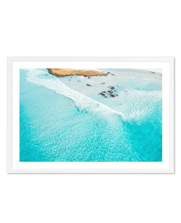 Aerial photograph of a coastal beach scene framed in white with white border. Turquoise ocean water transitions from deep blue-green in the foreground to lighter aquamarine near shore. White-capped waves roll onto pale sand beach dotted with dark rocks. Golden-brown rocky headland visible in upper portion. Composition captures the dramatic contrast between vibrant water colors and pristine sandy shoreline, with a serene, expansive mood conveying a sense of coastal tranquility.