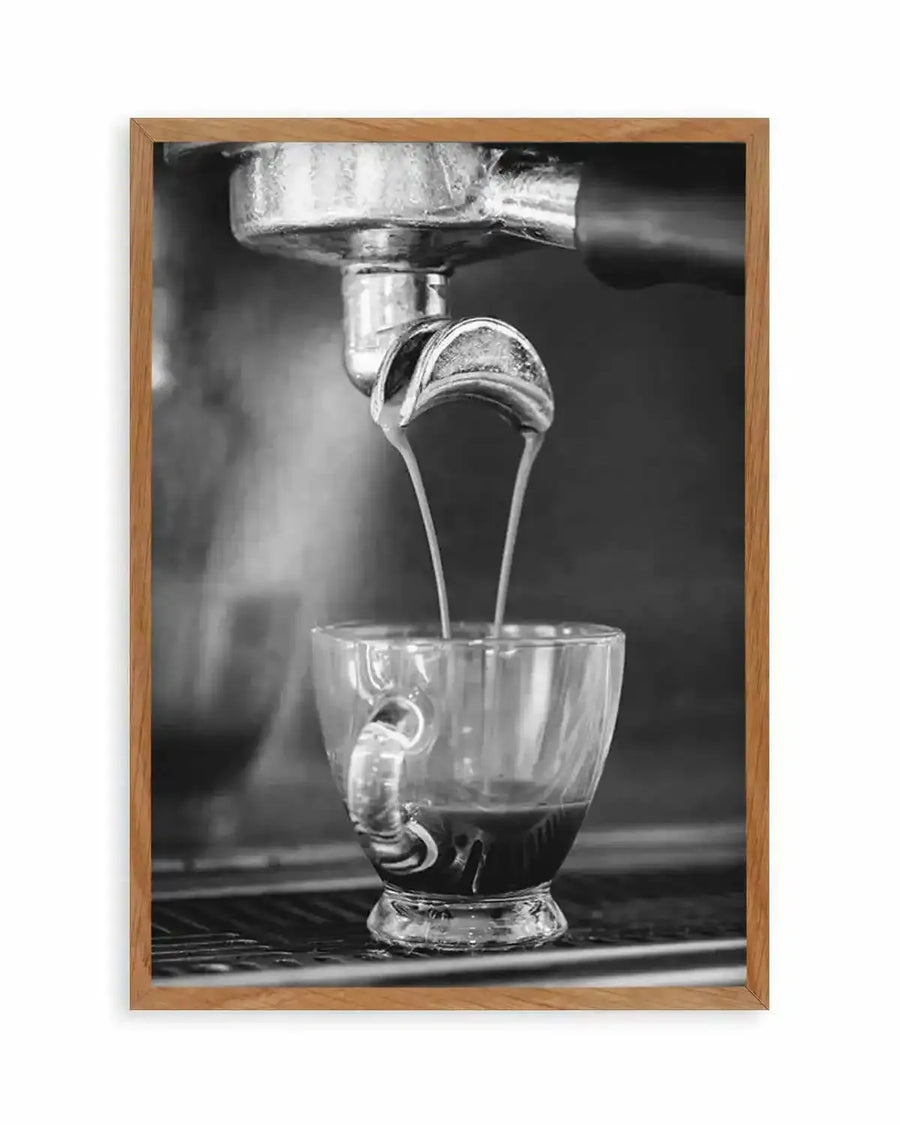 Framed black and white photograph of espresso being poured into a clear glass cup, with natural wood frame and white border. The composition captures two streams of espresso flowing from a portafilter into the cup, creating a rich dark liquid with a light crema on top. Steam rises from the cup, adding to the atmospheric mood.