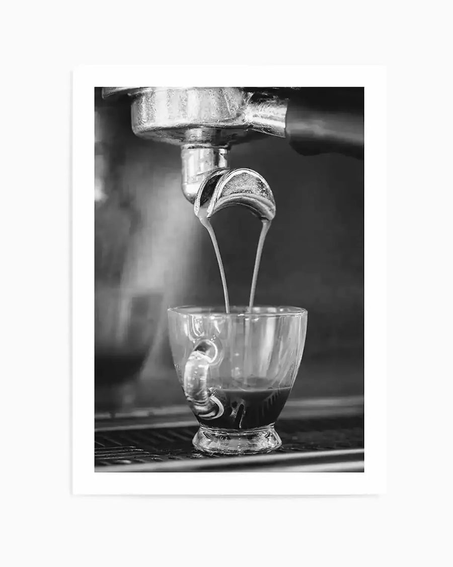 Framed black and white photograph of espresso being extracted from a portafilter into a clear glass cup, with a white border. Two streams of dark liquid pour from the portafilter into the cup, creating a rich crema.