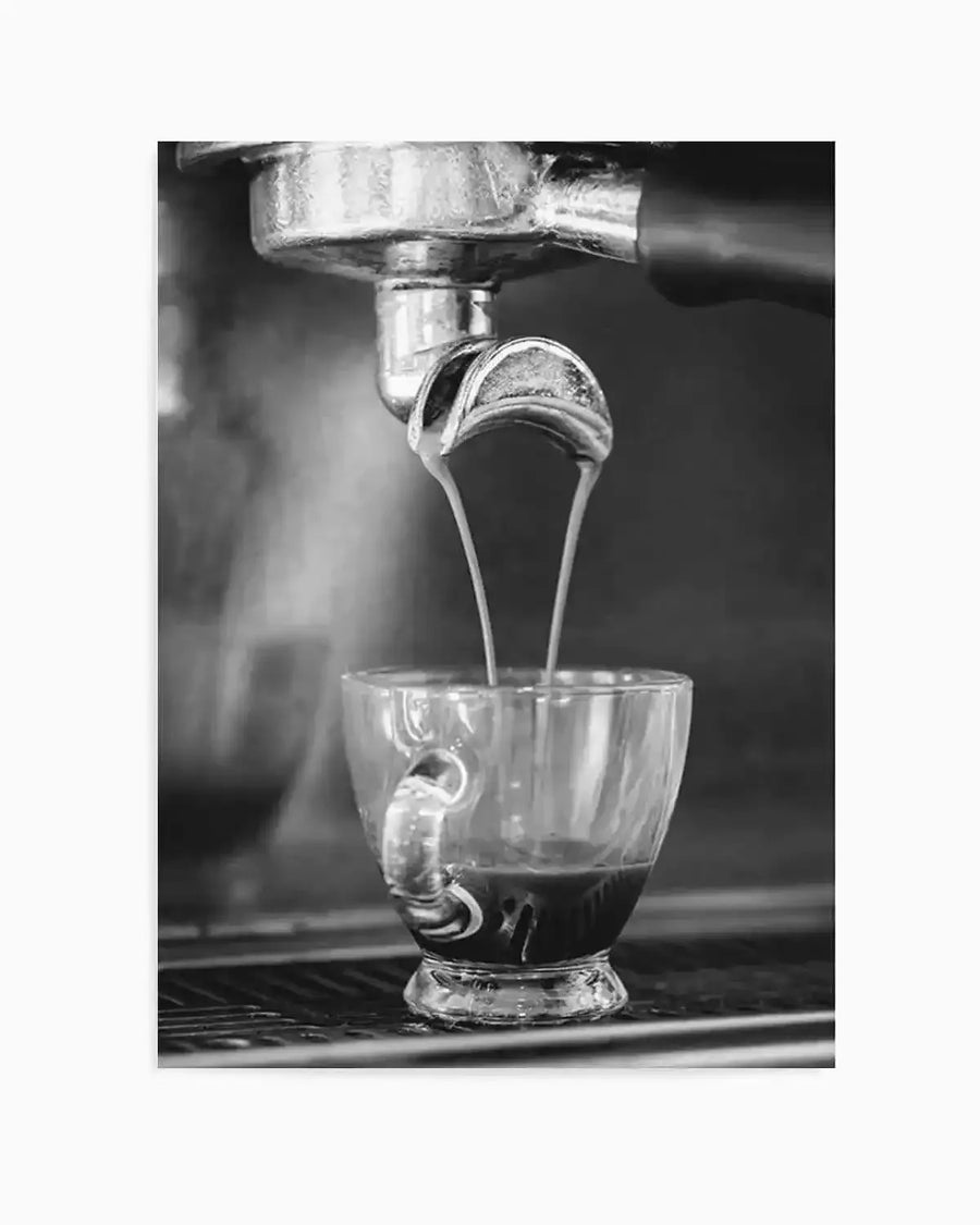 Black and white photograph art print capturing a close-up of an espresso machine pouring two streams of dark coffee into a clear glass cup. The metallic portafilter is visible at the top, with steam rising softly in the background. The composition highlights the rich texture of the coffee and the reflective surfaces of the machine.