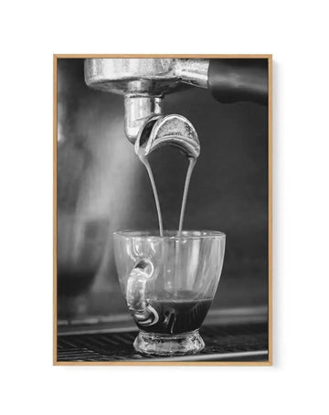 Black and white photograph of espresso pouring from a chrome espresso machine into a clear glass cup with decorative handle, captured mid-stream against a dark blurred background, mounted in a natural wood frame.