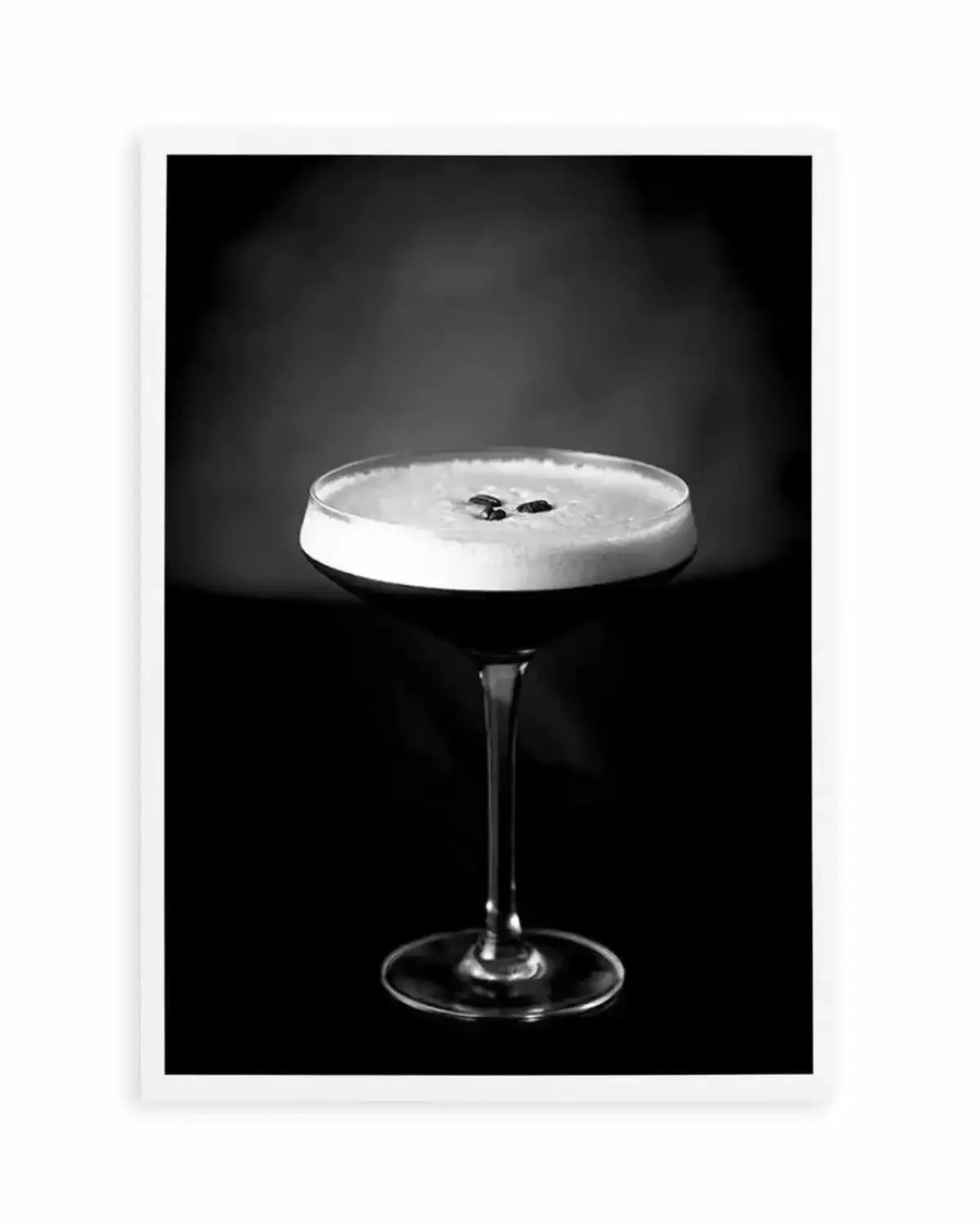 Black and white photograph of an Espresso Martini cocktail in a coupe glass against a dark background. The drink features a creamy white foam layer topped with two coffee beans. The glass has a thin stem and sits centered in the composition. The image is printed as an art print with a white border frame, creating a stark, minimalist aesthetic with dramatic lighting that emphasizes the contrast between the pale foam and the dark surroundings.