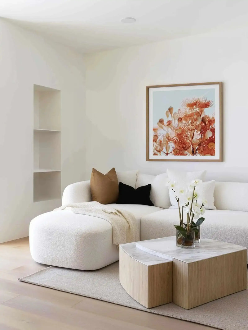 Square art print with wooden frame displayed on white bedroom wall above curved white sofa. Artwork features warm terracotta and rust-toned botanical illustration of flowering eucalyptus branches with delicate blooms against soft blue sky background. Print has white border within natural wood frame. Styled in minimalist bedroom with wooden cube side table, white throw pillow, potted white flowers, and light wood flooring.