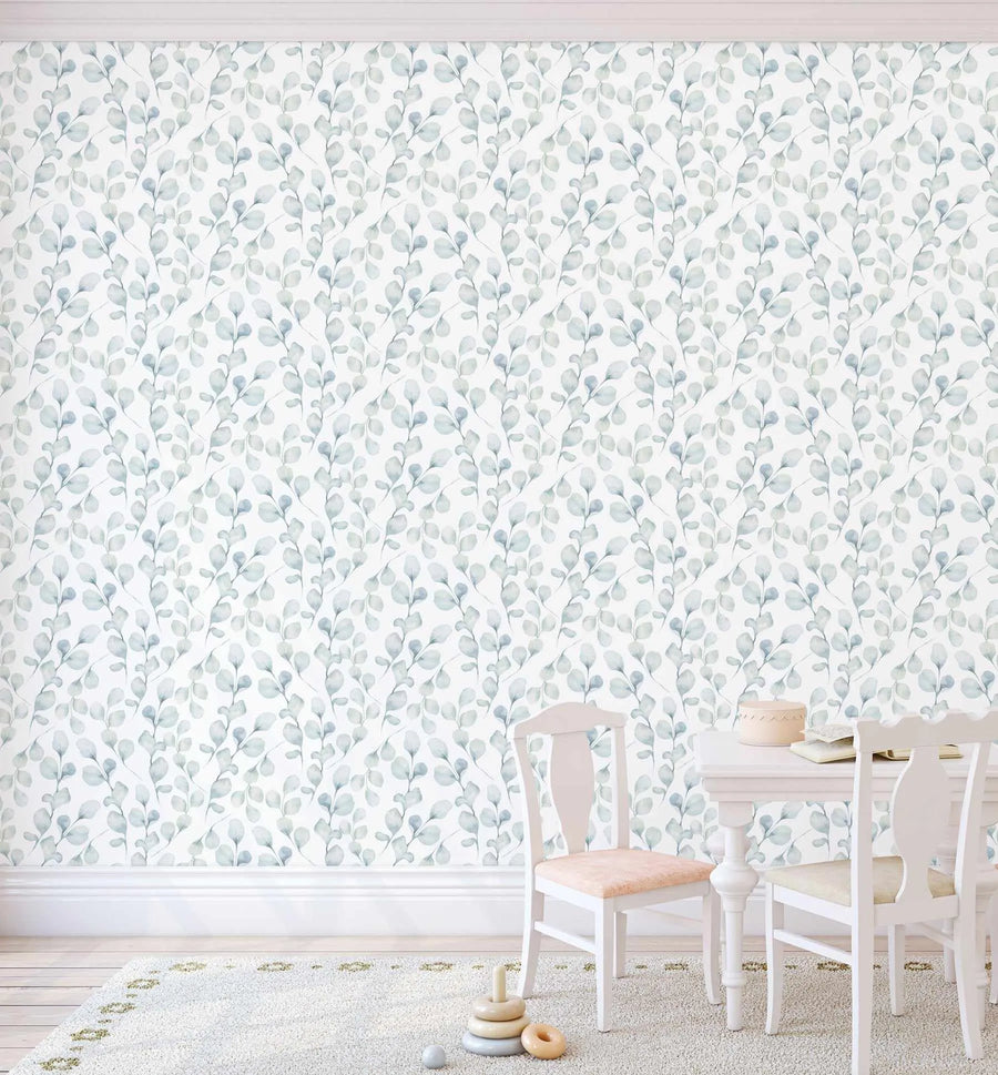 Peel and stick wallpaper featuring a repeating pattern of hand-painted eucalyptus branches in muted sage green and dusty blue on a soft white background. The botanical design has a loose, organic watercolour quality with visible brushstroke texture. The wallpaper is shown in a child's room above white wainscoting, with a small white table and chairs on a cream rug.
