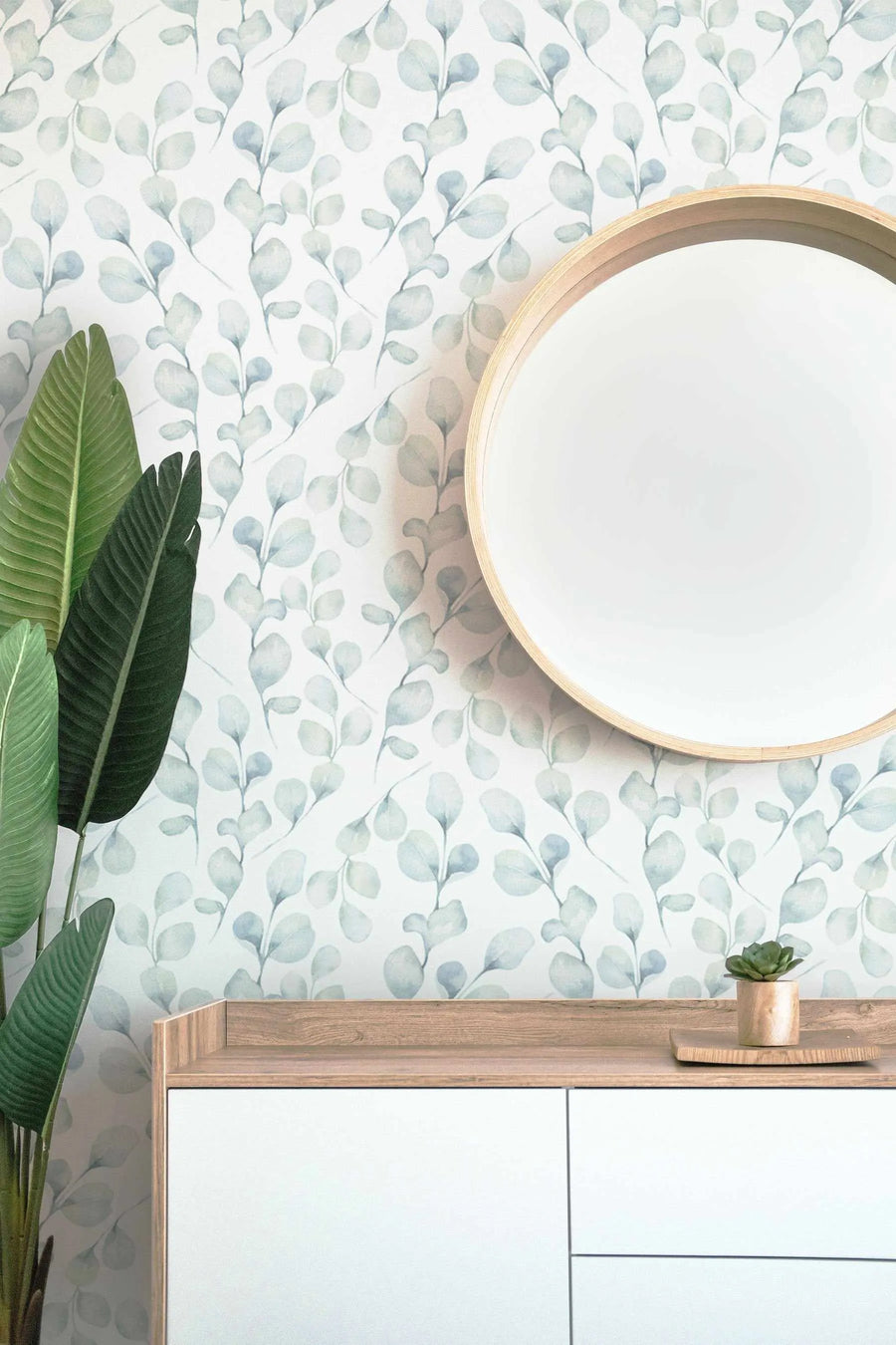 Lifestyle shot of a modern entryway featuring a round mirror with a light wood frame, a wooden console table with white drawers, and a large green plant. The wall is covered in peel and stick wallpaper with a repeating pattern of hand-painted eucalyptus branches in muted sage green and dusty blue on a soft white background. A small succulent in a wooden pot sits on the console table.