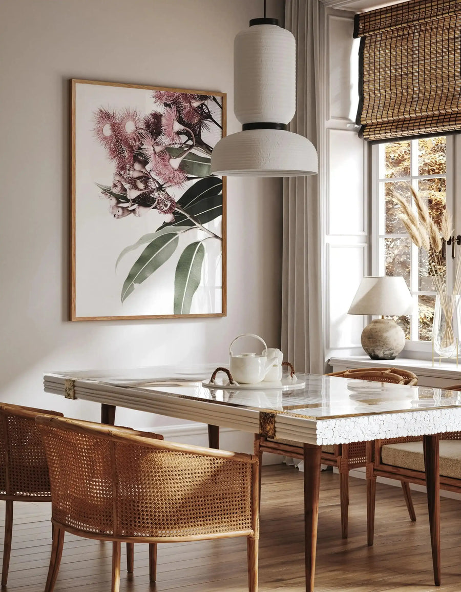 Eucalyptus in Bloom art print displayed on a cream wall above a marble-topped desk. The watercolor illustration features dusty rose and mauve flowers with dark green eucalyptus leaves against a white background, presented in a natural wood frame. The print hangs in a bright home office with rattan chairs, pendant lighting, and large windows with woven shades, creating a botanical, serene aesthetic.