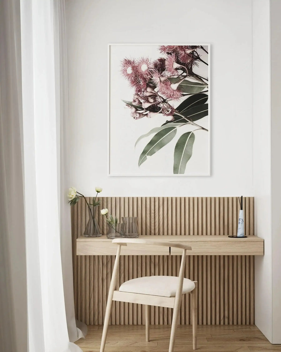 Home Office:
Framed art print of eucalyptus in bloom displayed above a light wood slatted desk in a minimalist home office. The print features a close-up photograph of pink eucalyptus flowers with spiky stamens and large green leaves against a white background, with a white frame and white border. The desk includes three clear glass vases with white flowers and a white marble diffuser.