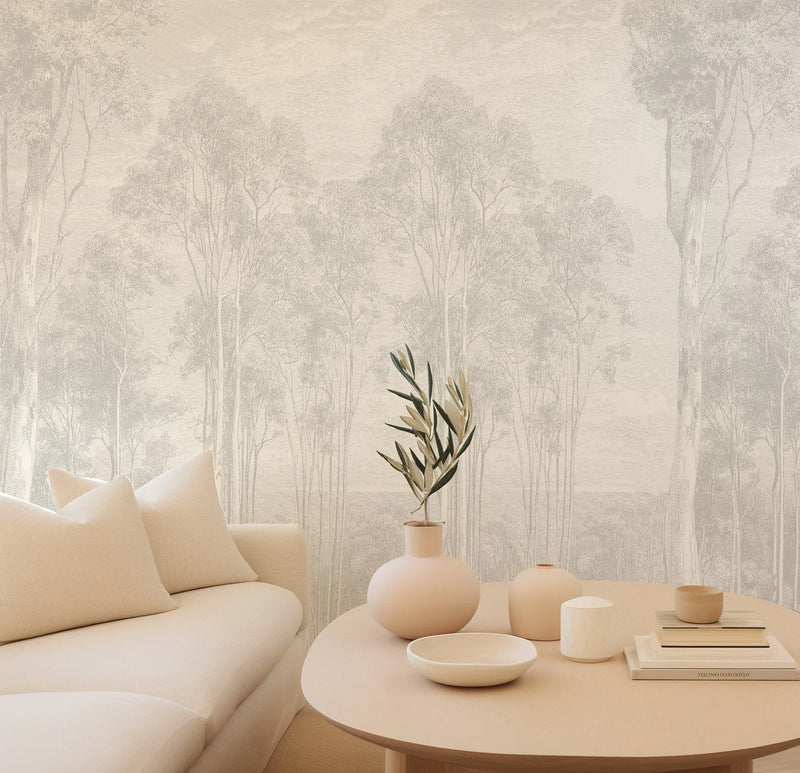 Lifestyle shot of a minimalist living room featuring a peel and stick wallpaper mural with a repeating pattern of hand-drawn eucalyptus trees in a pale grey on a warm cream background. The wallpaper creates a serene forest scene with varying tree heights and densities. A cream-colored sofa with textured cushions is visible on the left, and a light wood round coffee table with various decorative objects in muted tones is in the foreground.