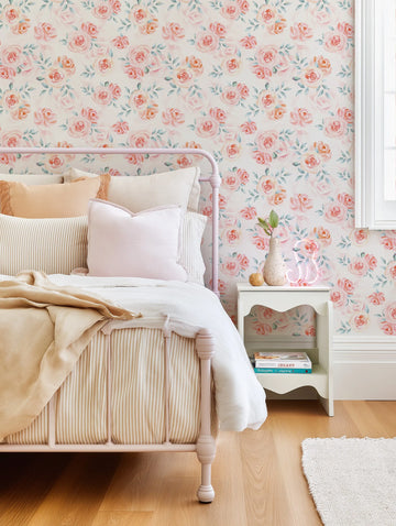 A bedroom featuring a light pink metal bed frame with striped beige and white bedding, a solid pink pillow, and a textured beige throw. The wall behind the bed is adorned with wallpaper featuring a repeating pattern of hand-painted watercolor roses in shades of pink and peach with soft green leaves on a cream background. A white nightstand with books and a vase with greenery sits to the right of the bed.