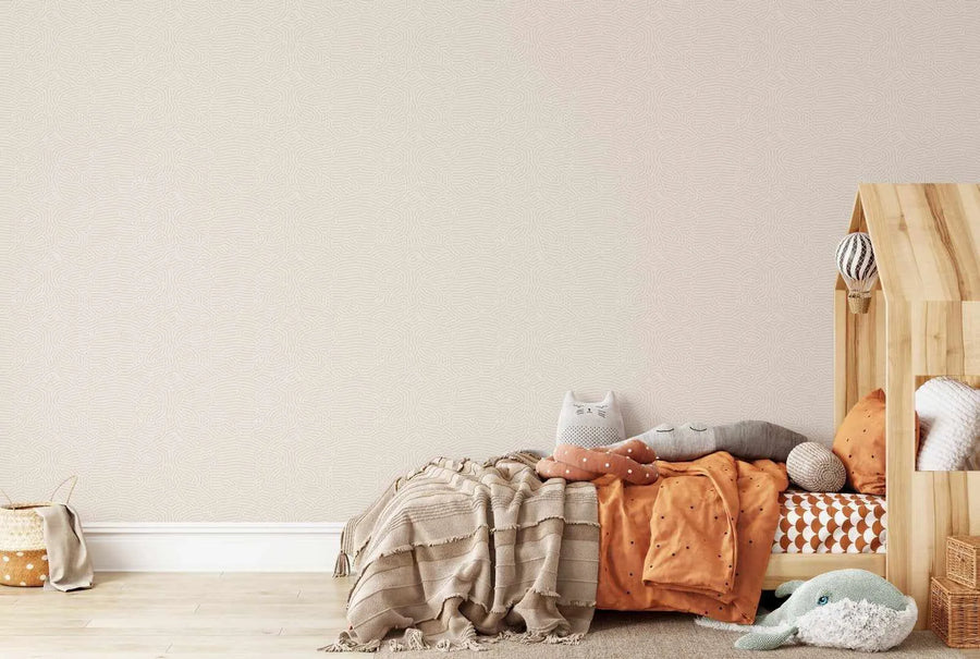 Lifestyle shot of a child's bedroom featuring Eyre Wallpaper. The wallpaper has a repeating pattern of organic, flowing lines in a light beige tone on a creamy white background, creating a subtle, textured appearance. The room includes a wooden house-shaped bed with orange bedding, a striped blanket, and various plush toys, including a whale, a cat, and a cloud. A woven basket with a beige cloth sits on the light wood floor in the foreground.