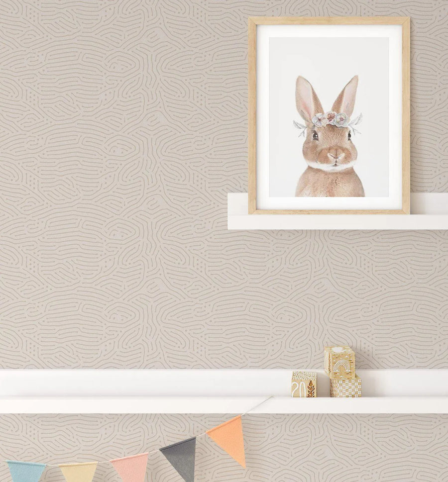Nursery:
Eyre Wallpaper featuring a repeating pattern of thin, organic, squiggly lines in a light grey tone on a warm, light beige background, creating a subtle, textured effect. The wallpaper is visible on the wall of a nursery, with a white shelf displaying a framed art print of a brown bunny wearing a floral wreath on its head. Below, another white shelf holds three wooden blocks, and a string of pastel-coloured bunting hangs across the bottom.