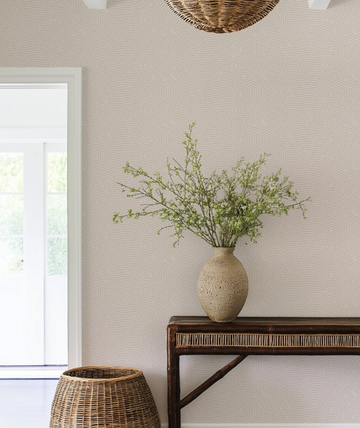Lifestyle shot of a hallway featuring Eyre Wallpaper with a subtle, organic pattern of thin, wavy lines in a light beige tone on a slightly lighter cream background. The wallpaper is visible on the right wall, behind a dark wooden console table with woven detailing, a textured light brown vase with green branches, and a large woven basket on the floor. A white door frame is visible on the left.