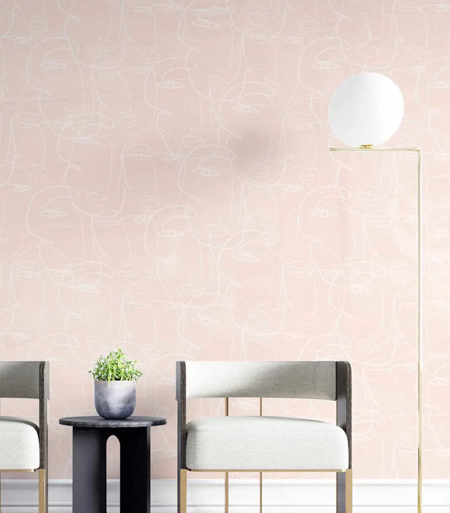 Peel and stick wallpaper featuring a repeating pattern of abstract faces in continuous line art style, rendered in off-white against a soft blush pink background. The design has a minimalist, hand-drawn quality. The wallpaper is shown in a room with two modern chairs and a side table with a plant.