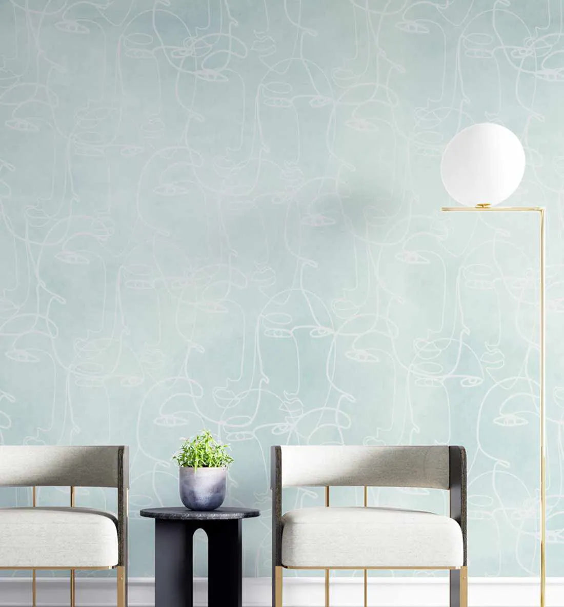 Lifestyle shot of a room featuring peel and stick wallpaper with a repeating pattern of abstract faces in white line art on a seafoam green background. The wallpaper covers a wall behind two modern chairs with light grey upholstery and gold frames, flanking a small black side table with a potted plant. A tall gold floor lamp with a spherical white shade stands on the right.