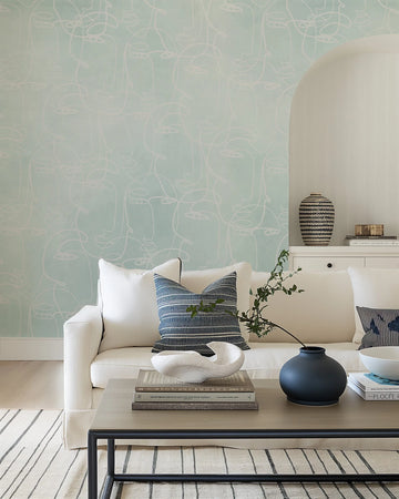 Lifestyle shot of a minimalist living room featuring peel and stick wallpaper with a repeating pattern of abstract line art faces in white on a seafoam green background. The wallpaper covers a large wall behind a white sofa with blue and white striped cushions. A light wood coffee table with books and decorative objects sits in front of the sofa.