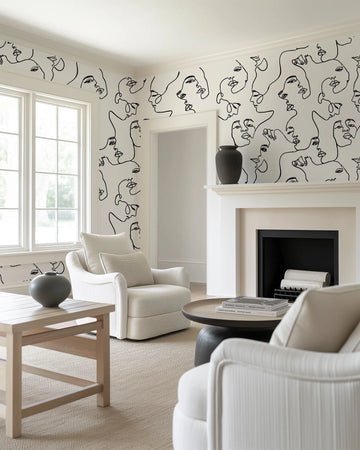Lifestyle shot of a minimalist living room featuring Faces peel and stick wallpaper. The wallpaper has a repeating pattern of abstract, continuous line art faces in black on a white background, covering the walls around large windows and a fireplace. The room includes two cream upholstered armchairs, a light wood coffee table with a dark grey vase, and a dark wood round side table with books.