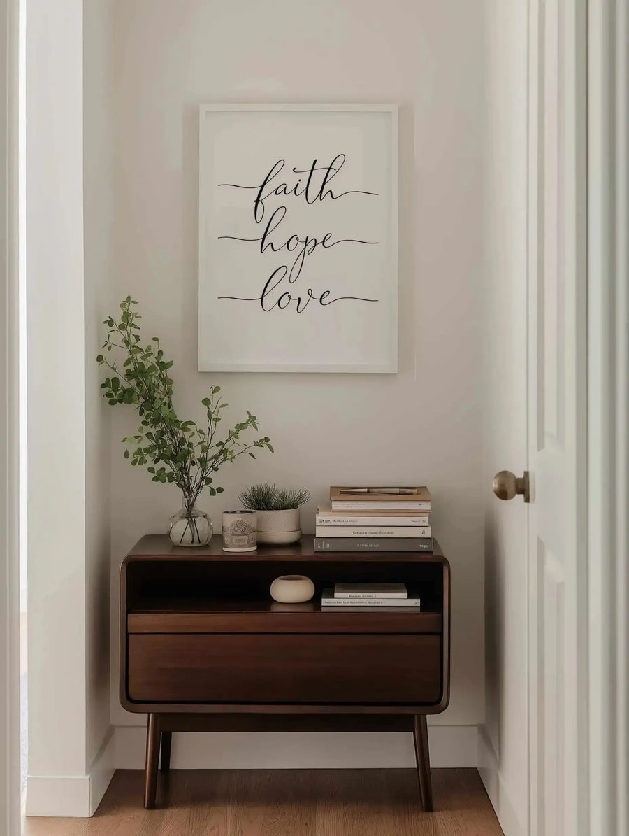 Hallway:
Framed art print displayed above a dark wood console table in a neutral-toned hallway. The print features the words "faith hope love" in elegant black script on a white background, with a white frame. The console table is styled with a clear vase of green leafy branches, a small potted plant, a candle, and stacks of books. A white door with a brass knob is visible on the right.