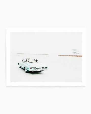 Framed photograph of a light blue vintage convertible car with two people inside, driving on a wide, empty, light-colored road or beach. The car is seen from the rear, slightly to the left of the frame, with a vast, bright, almost white background and faint horizontal lines suggesting a distant horizon or structures.