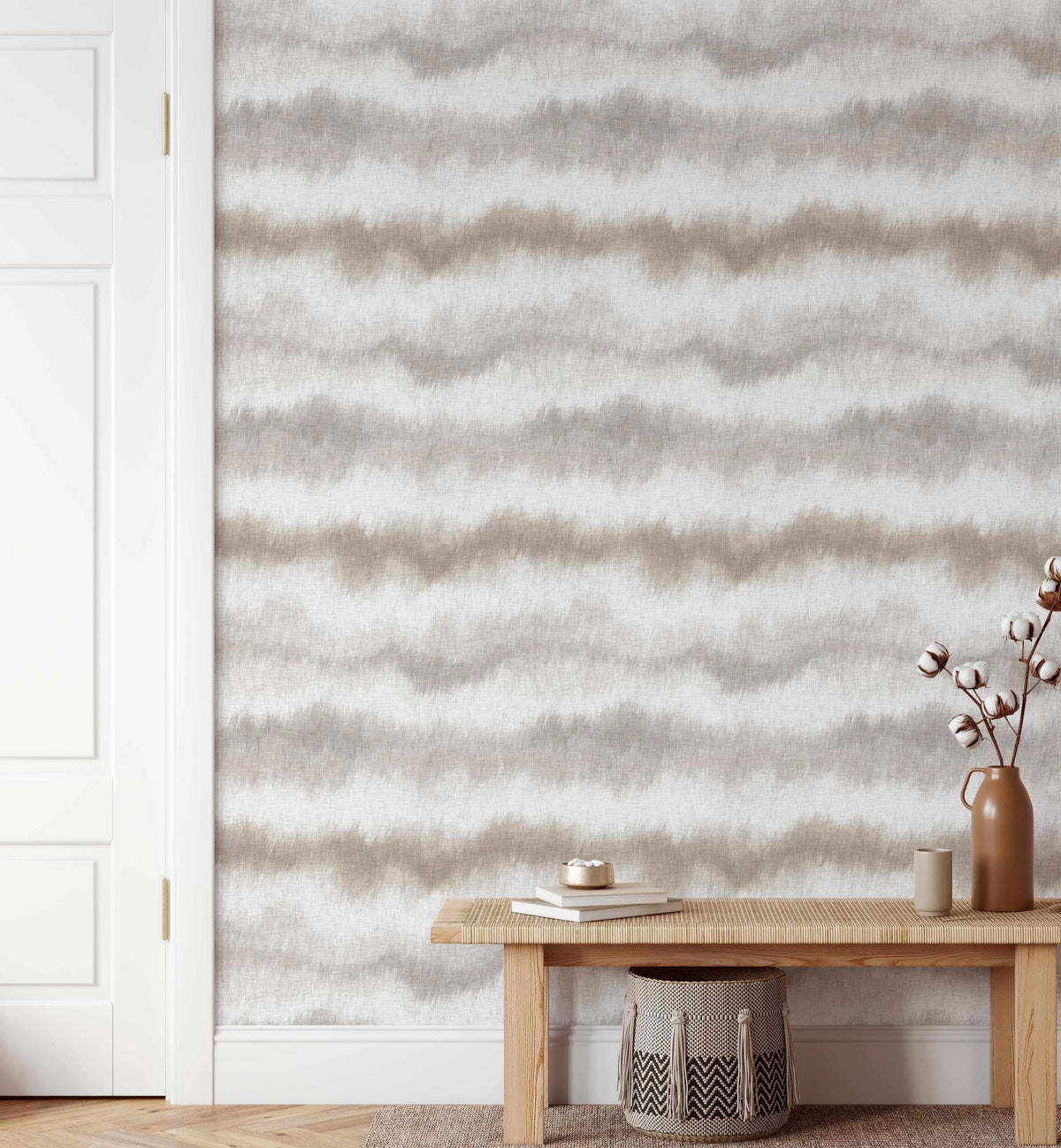 Lifestyle shot of a room with faux linen wallpaper featuring horizontal, undulating stripes in varying shades of light beige, taupe, and off-white, creating a textured, organic pattern. The wallpaper covers a wall next to a white door with gold hardware. A light wood bench with a woven top sits against the wall, holding a brown ceramic vase with cotton branches, a small gold bowl, and two books. A woven basket with a black and white chevron pattern is placed under the bench.