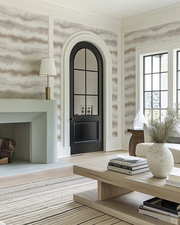 Lifestyle shot of a living room featuring faux linen wallpaper with a repeating horizontal stripe pattern in muted taupe and off-white, creating a soft, undulating effect. The wallpaper covers the upper walls, framed by white crown molding and trim around an arched black door and two large windows with black grilles. A light green fireplace with a lamp on the mantel is visible on the left, and a light wood coffee table with books and a textured white vase with dried foliage is in the foreground.