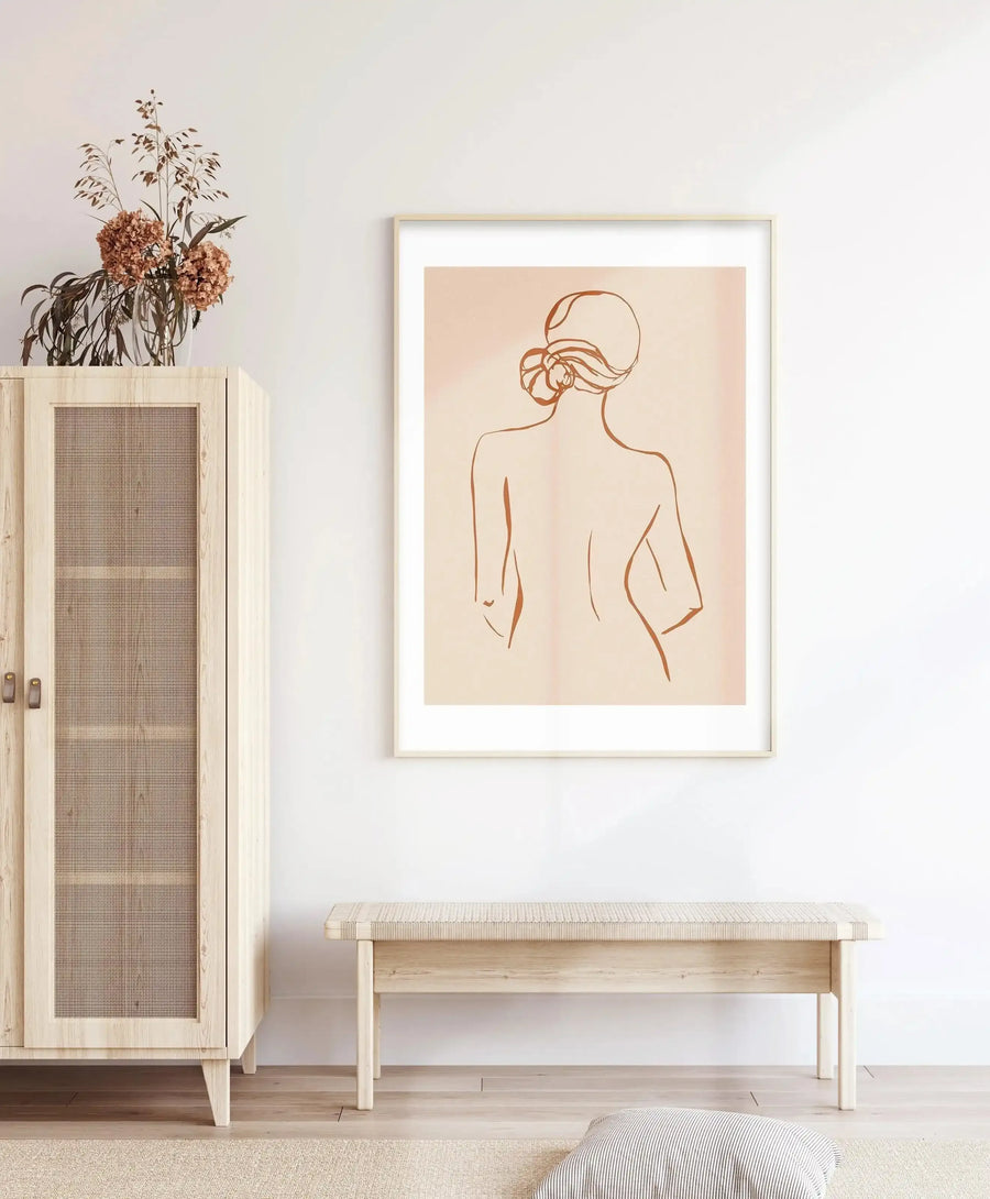 Lifestyle shot featuring a framed art print displayed above a light wood bench in a minimalist entryway. The print depicts a minimalist line art illustration of a woman's back and head with hair in a bun, rendered in terracotta on a soft peach background, with a natural oak frame and white border. To the left, a light wood cabinet with a woven cane door holds a vase of dried flowers. A striped cushion rests on a cream rug in the foreground.