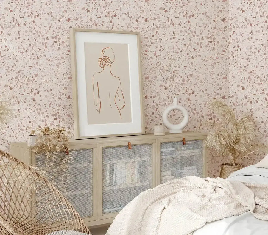 Framed art print of a female form displayed on a light wood cabinet in a bedroom with terrazzo wallpaper. The print features a minimalist line drawing of a woman's back with her hair in an updo, in a terracotta hue on a cream background, with a natural wood frame and white border. The room includes a wicker chair, a textured cream throw on a bed, and dried pampas grass in a white ceramic vase.