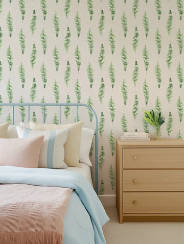 Lifestyle shot of a bedroom featuring Fern Foliage Wallpaper with a repeating pattern of hand-painted fern fronds in varying shades of green on a light cream background. The wallpaper is visible behind a light blue metal bed frame with a bed dressed in light blue and pink bedding, and an assortment of pillows in cream, light blue, and yellow patterns. To the right of the bed is a light wood bedside table with three drawers, on which sits a stack of books and a clear vase with green foliage.