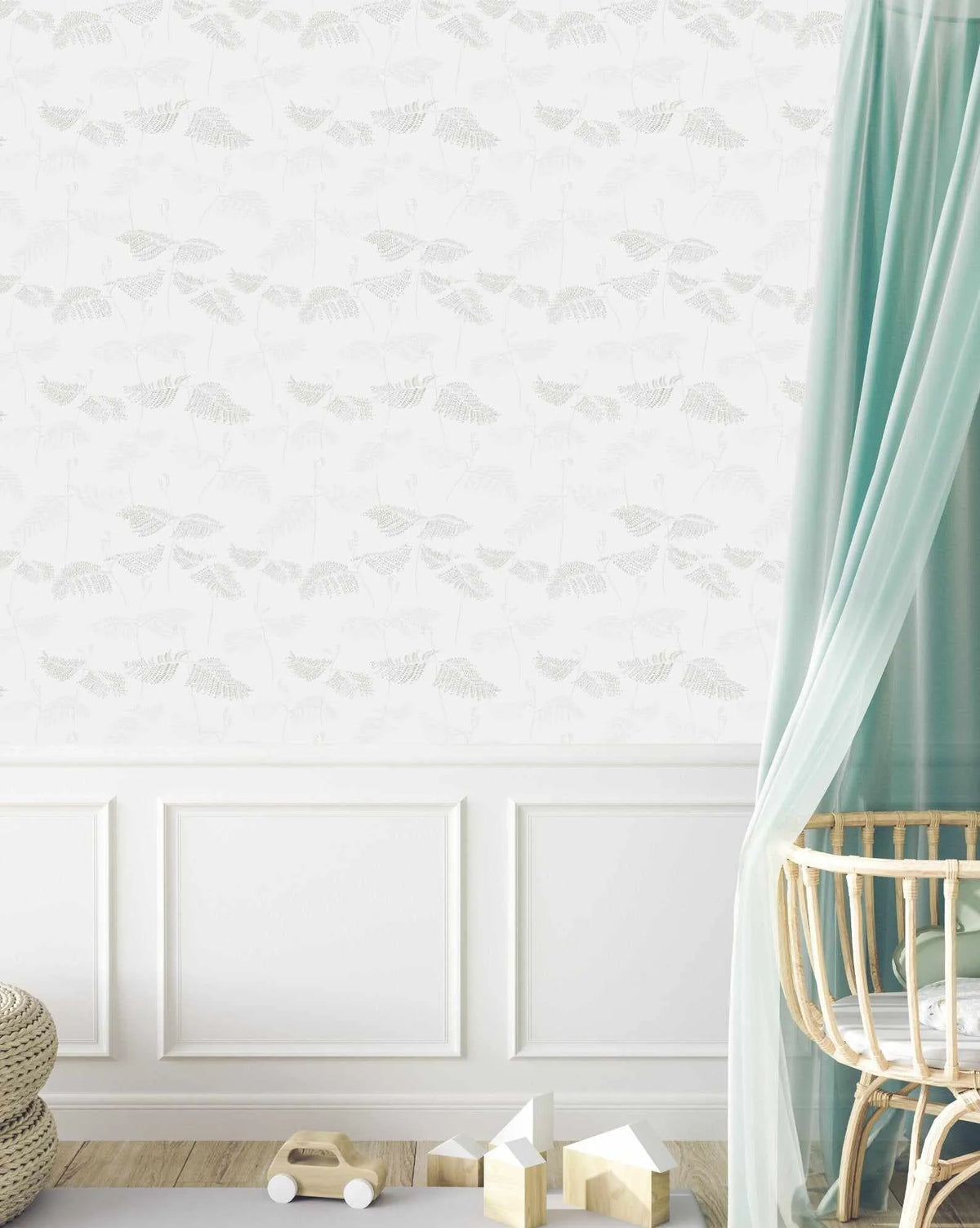 Lifestyle shot of a nursery featuring a wall covered in "Ferns in Grey" wallpaper. The wallpaper has a repeating pattern of delicate fern leaves in a subtle light grey tone on a white background, creating a soft, textural effect. The lower third of the wall is white wainscoting with rectangular paneling. To the right, a light blue sheer curtain hangs over a natural rattan baby crib. On the light wood floor, there's a woven basket, a wooden toy car, and several small wooden house blocks.