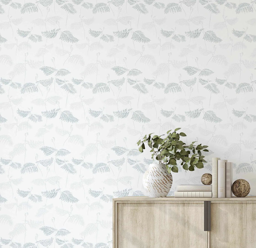 Ferns in Seafoam & Moss Wallpaper displayed on a wall behind a light wood cabinet. The wallpaper features a repeating pattern of delicate fern fronds in seafoam green and muted moss tones illustrated against a soft cream background. The design has a botanical, hand-drawn quality with fine line work and subtle texture. A white ceramic vase with green foliage, stacked cream-colored books, and decorative woven spheres are styled on the cabinet shelf in front of the wallpaper.