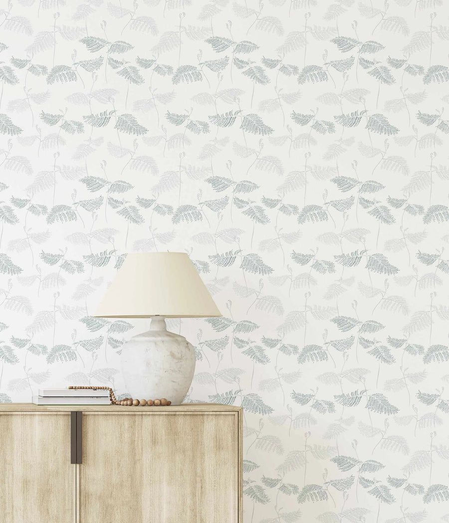 "Peel and stick wallpaper featuring a repeating pattern of delicate fern fronds in muted seafoam green and soft moss green, with some fronds in a lighter grey-green, on a clean white background. The botanical design has a hand-drawn, illustrative quality with fine line details."