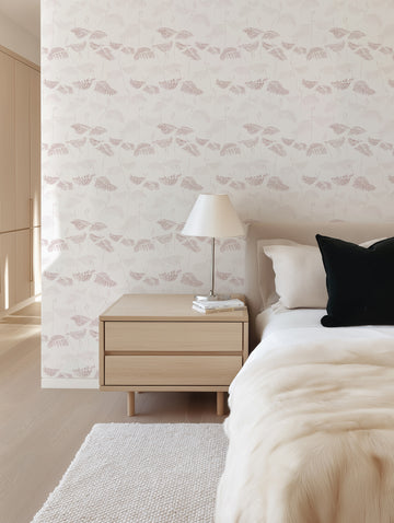 Lifestyle shot of a bedroom featuring Ferns in Burgundy & Beige wallpaper on the accent wall behind the bed. The wallpaper has a repeating pattern of delicate fern fronds in varying shades of burgundy and beige on a light cream background, creating a subtle botanical design. The room includes a light wood bedside table with a lamp and books, a bed with white and black pillows, and a cream faux fur throw, all contributing to a soft and inviting atmosphere.