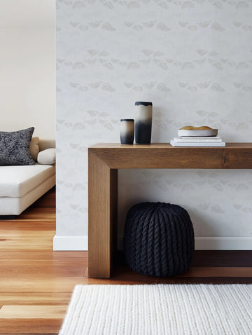 "Ferns in Grey wallpaper featuring a repeating pattern of delicate fern fronds in a subtle grey on a light grey background, creating a soft, textured look. The wallpaper is displayed on a wall behind a wooden console table with two ceramic vases and books, and a dark grey knitted pouf underneath. A cream sofa with a patterned cushion is visible in the foreground to the left."