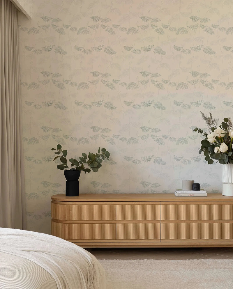 Ferns in Seafoam & Moss Wallpaper displayed on a bedroom wall above a wooden dresser. The wallpaper features a repeating pattern of delicate fern fronds in soft seafoam green and muted moss tones on a warm cream background. The illustrated botanical design has a subtle, naturalistic quality with fine linework. The room includes a wooden dresser with ribbed detailing, a black vase with eucalyptus, a white ceramic vase with white flowers, cream curtains, and a white bedding corner visible in the foreground.