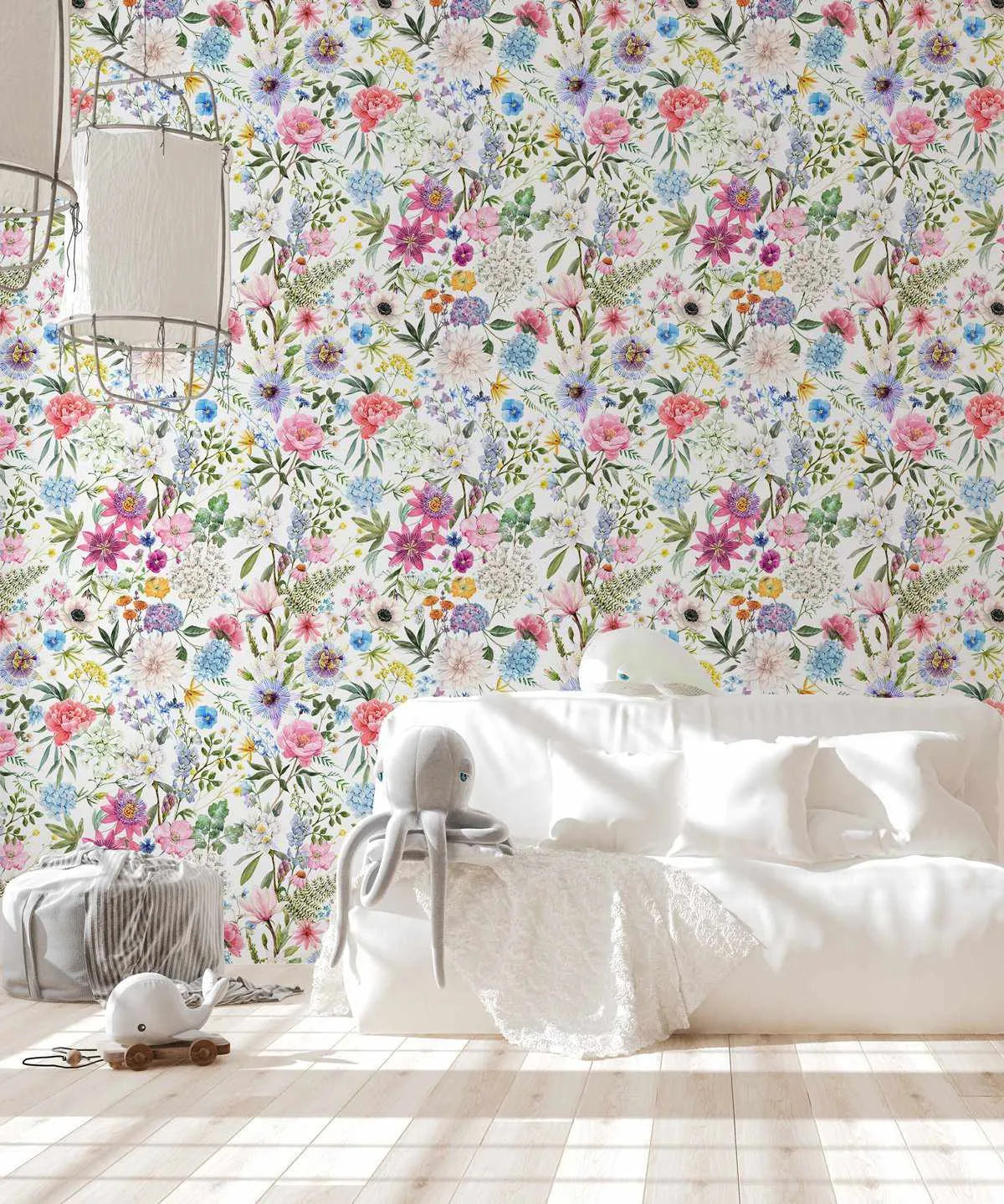 Nursery:
"Peel and stick wallpaper featuring a repeating pattern of vibrant, hand-painted wildflowers in various shades of pink, blue, purple, yellow, and white on a crisp white background, displayed in a bright nursery. The botanical design has a loose, organic watercolor quality with visible brushstroke texture. The room includes a white sofa with white pillows and a white lace throw, a grey octopus plush toy, a grey striped pouf, and a white whale pull toy on a light wood floor."