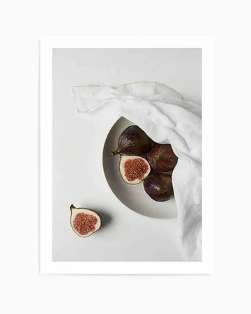 "Photography art print featuring a top-down view of fresh figs on a white surface. A white linen cloth drapes over a white bowl containing several whole, dark purple figs. One fig is cut in half, revealing its vibrant red-pink flesh and tiny seeds. Another half-fig lies outside the bowl on the white surface, mirroring the internal colours."