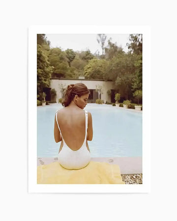 Framed colour photograph of a woman sitting by a swimming pool, viewed from behind. The woman has dark hair tied back and is wearing a white, low-backed swimsuit, sitting on a yellow towel at the edge of a light blue pool. In the background, lush green trees and a light-coloured building with potted plants are visible under an overcast sky.